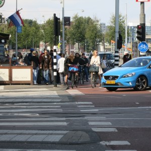 Street chaos. On the left, the cafe has taken 75% of the sidewalk for seating. In the middle, bikes spill out of the cycle lane, vying for who gets to cross first. Pedestrians are squeezed into a couple feet. Tram tracks down the middle of the street.